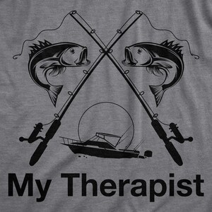 May include: A gray t-shirt with a black graphic design of two crossed fishing rods with fish on the lines and a boat in the middle. The text "My Therapist" is printed below the graphic.