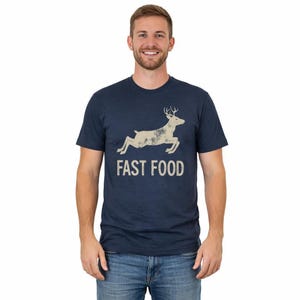 May include: A navy blue short-sleeve t-shirt featuring a beige graphic of a leaping deer above the text "FAST FOOD" in a distressed, sans-serif font. The shirt has a crew neckline and a standard fit, showcasing a clean and simple design.