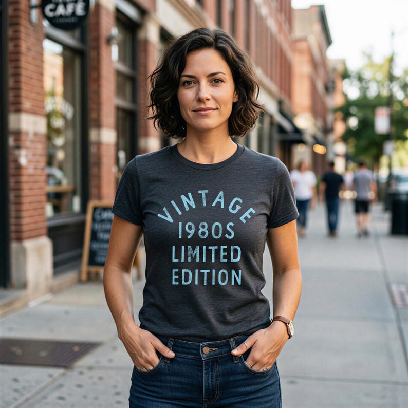 May include: A woman wearing a dark gray t-shirt with the text "VINTAGE 1980S LIMITED EDITION" in light blue. She is wearing blue jeans and a brown watch. The background shows a brick building and a sidewalk.
