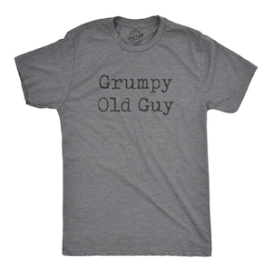 May include: A heather grey short-sleeve crew neck t-shirt featuring the text Grumpy Old Guy printed in a dark charcoal typewriter-style font across the chest. The shirt has a standard fit with a ribbed collar and a visible brand label inside.