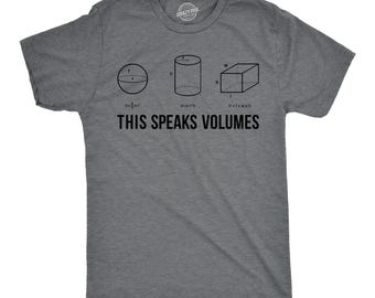 May include: A charcoal gray short-sleeve t-shirt featuring black line drawings of a sphere, a cylinder, and a rectangular prism with their respective volume formulas printed below each shape. The text THIS SPEAKS VOLUMES is printed in black.