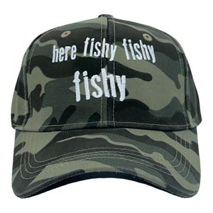 May include: A camouflage baseball cap featuring the embroidered white text here fishy fishy fishy on the front panel. The hat has a structured crown, a curved brim, and small circular ventilation eyelets on the top panels.