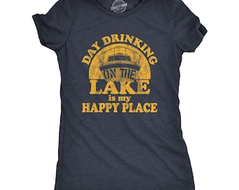 Op de afbeelding: Een donkerblauw t-shirt met korte mouwen en een verweerde gele afbeelding van een boot op het water. De tekst luidt "DAY DRINKING ON THE LAKE is my HAPPY PLACE" in een vet, versleten lettertype. Het shirt heeft een ronde halslijn.