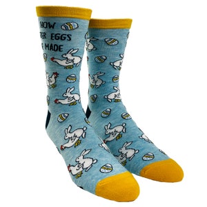 Peut inclure: Une paire de chaussettes bleues avec un revers jaune. Les chaussettes présentent un motif répétitif de lapins blancs et d'œufs bleus. Le texte "How Easter Eggs Are Made" est imprimé sur les chaussettes.