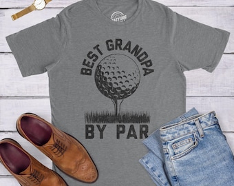 Puede incluir: Una camiseta gris jaspeado de manga corta con un gráfico negro de una pelota de golf sobre un tee y el texto BEST GRANDPA BY PAR. La camiseta se muestra junto a un reloj de pulsera dorado con correa de cuero marrón y vaqueros azules.