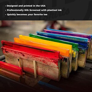 May include: A close-up of several screen printing squeegees with vibrant colors of red, orange, yellow, green, blue, and purple. The text reads: Designed and printed in the USA, Professionally Silk Screened with plastisol ink, Quickly becomes your favorite tee.
