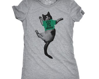 May include: A heather gray short-sleeve t-shirt featuring a black and white cat wearing a green shirt with the text PET ME I'M IRISH printed in black. The cat is depicted in a dancing pose with one leg raised and arms extended outward.