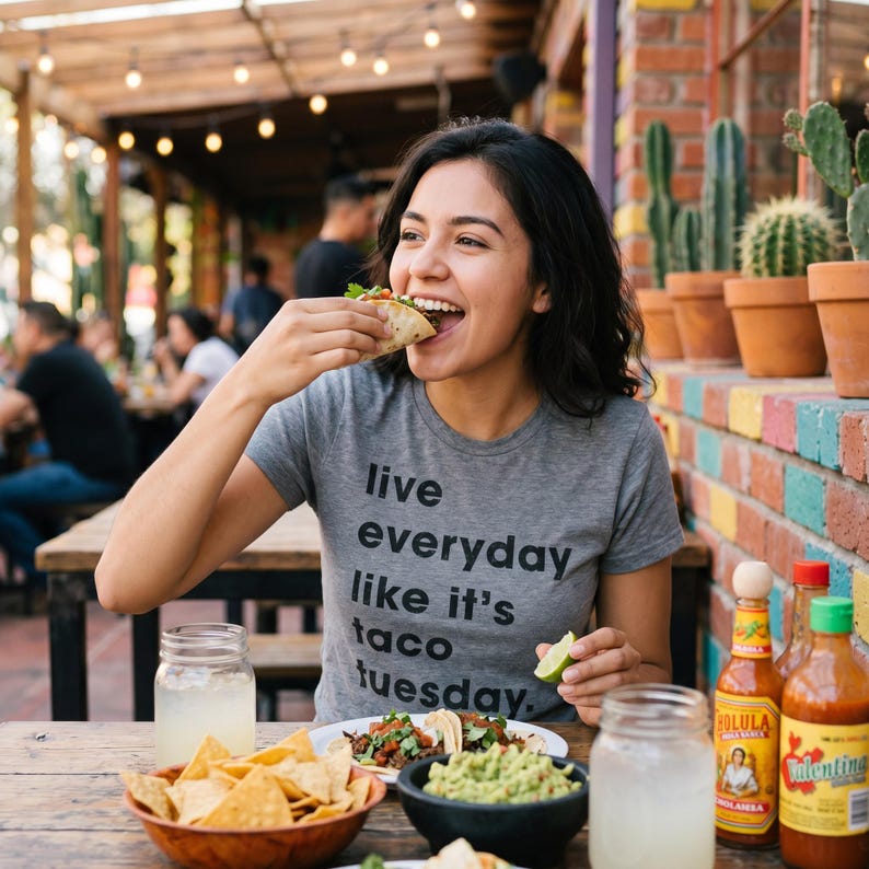 May include: A person wearing a grey t-shirt with the text "Live everyday like it's taco tuesday" is eating a taco. A table is set with chips, guacamole, tacos, and drinks. Bottles of hot sauce are visible.
