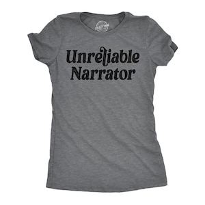May include: A charcoal grey short-sleeved t-shirt featuring the text "Unreliable Narrator" printed in black across the chest. The shirt has a crew neckline and a small "Crazy Dog" logo printed on the inside of the collar area.