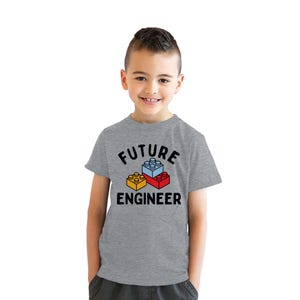 May include: A gray t-shirt with the words "FUTURE ENGINEER" in black, along with a graphic of three colorful toy blocks. The blocks are yellow, blue, and red. The shirt is short-sleeved.