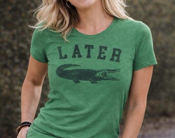 May include: A green short-sleeved t-shirt featuring the word LATER printed in a dark, distressed font above a graphic illustration of an alligator. The shirt has a crew neckline and is made of a heathered fabric with a textured appearance.