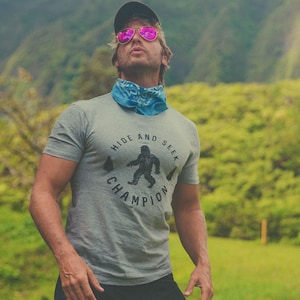 May include: A man wearing a grey t-shirt with a black and white graphic of a Bigfoot. The graphic says "Hide and Seek Champion". He is wearing pink sunglasses and a blue bandana.