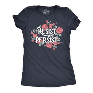 Op de afbeelding: Een donker antracietgrijs t-shirt met korte mouwen en een grafische print van roze rozen met groene bladeren rondom de witte tekst "RESIST AND PERSIST". Het shirt heeft een ronde hals en een gemêleerde textuur.
