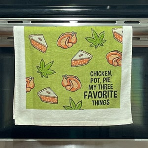 May include: A white kitchen towel with a green background and a repeating pattern of chicken, pie, and marijuana leaves. The text "Chicken, Pot, Pie, My Three Favorite Things" is printed in black on the towel.