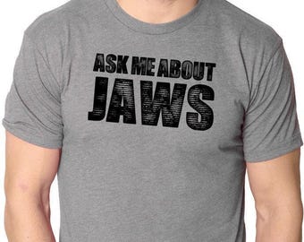 May include: A heather gray short-sleeved crew neck t-shirt featuring the black text "ASK ME ABOUT JAWS" printed across the chest. The shirt is made of a soft, lightweight fabric with a standard fit, short sleeves, and a classic round neckline.