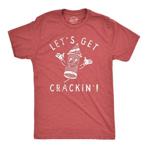 May include: A heather red short-sleeve t-shirt featuring a white distressed graphic of a cartoon firework character with arms and a face. The text above and below the firework reads LET'S GET CRACKIN'! in a distressed white font style.