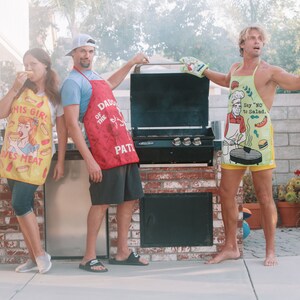 Op de afbeelding: Drie mensen dragen schorten met humoristische uitspraken over eten en staan voor een zwarte barbecue. De persoon links draagt een gele schort met de tekst "Dit meisje houdt van vlees". De persoon in het midden draagt een rode schort met de tekst "Daddy of the grill patrol". De persoon rechts draagt een groene schort met de tekst "Zegt 'nee' tegen salade."