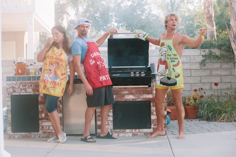 Op de afbeelding: Drie mensen dragen schorten met humoristische uitspraken over eten en staan voor een zwarte barbecue. De persoon links draagt een gele schort met de tekst "Dit meisje houdt van vlees". De persoon in het midden draagt een rode schort met de tekst "Daddy of the grill patrol". De persoon rechts draagt een groene schort met de tekst "Zegt 'nee' tegen salade."