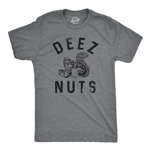 May include: A heather grey short-sleeve t-shirt featuring a black graphic of a squirrel holding two acorns. The text DEEZ NUTS is printed in black capital letters above and below the squirrel illustration. The shirt has a crew neckline.
