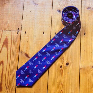 May include: A dark blue necktie featuring a repeating pattern of red, white, and blue rocket-shaped ice pops. Each ice pop graphic is accompanied by the text "COOLEST POP" printed in white letters. The tie is rolled at the top end.