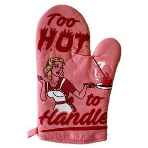 May include: A pink oven mitt featuring a graphic illustration of a person with blonde hair wearing an apron and holding a frying pan with a flame. The text "Too HOT to Handle" is printed in bold red lettering across the top and bottom sections.