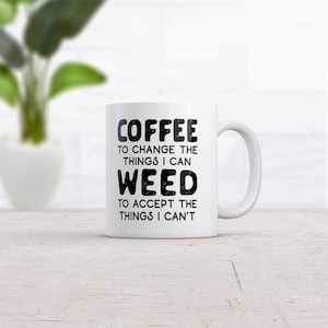 May include: A white ceramic mug with a handle featuring black text that reads: COFFEE TO CHANGE THE THINGS I CAN WEED TO ACCEPT THE THINGS I CAN'T. The mug has a smooth finish and a cylindrical shape, displaying bold, distressed-style lettering.
