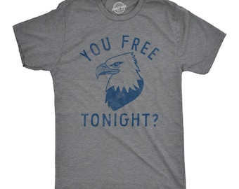 Puede incluir: Una camiseta gris carbón de manga corta con un gráfico azul desgastado de una cabeza de águila calva en el centro. El texto "YOU FREE TONIGHT?" está impreso en letras mayúsculas azules, con "YOU FREE" sobre el águila y "TONIGHT?" debajo.