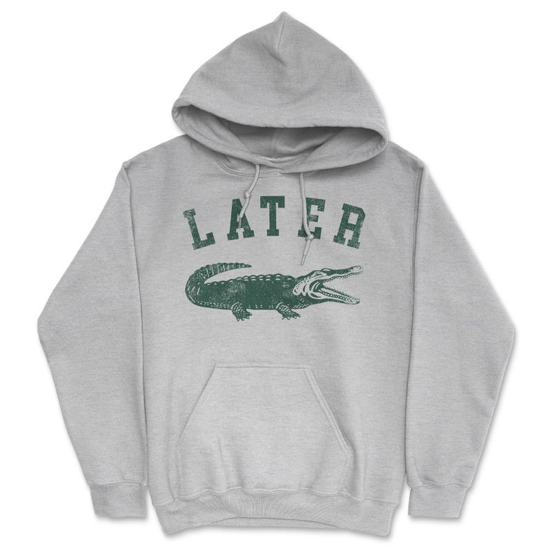 Puede incluir: Una sudadera con capucha gris con la palabra "LATER" impresa en verde en la parte delantera. Debajo de la palabra hay un gr&aacute;fico de un cocodrilo verde.