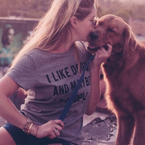May include: A gray t-shirt with the text "I LIKE DOGS AND MAYBE PEOPLE" is worn by a person kissing a golden retriever. The dog is on a leash. The image has a soft, warm color palette.