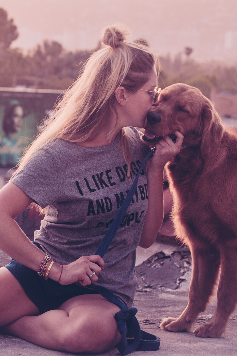 May include: A gray t-shirt with the text "I LIKE DOGS AND MAYBE PEOPLE" is worn by a person kissing a golden retriever. The dog is on a leash. The image has a soft, warm color palette.