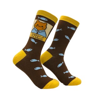 Könnte beinhalten: Ein Paar braune Socken mit gelben Akzenten. Eine Socke zeigt eine Cartoon-Katze mit blauem Hemd und dem Text "Employee of the Month". Beide Socken haben hellblaue Fischmuster. Die Socken sind für Komfort und Stil konzipiert.