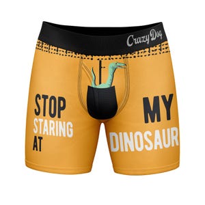 Può includere: Boxer arancioni con elastico in vita nero con la scritta Crazy Dog. La parte anteriore presenta una grafica a tasca nera con una testa di dinosauro verde chiaro che ne esce, insieme al testo STOP STARING AT MY DINOSAUR.
