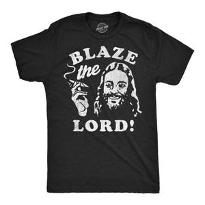 May include: Black t-shirt with a white graphic of a man with long hair and a beard holding a joint. The text on the shirt reads "Blaze the Lord!"