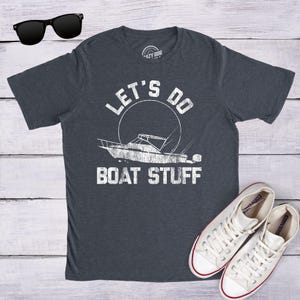May include: A dark heather grey short-sleeve t-shirt featuring a white distressed graphic of a boat inside a circle with the text "LET'S DO BOAT STUFF" printed above and below the boat. Black sunglasses and white canvas trainers are also visible.