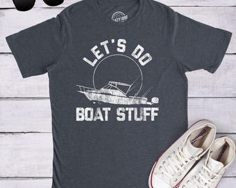 Op de afbeelding: Een donkergrijs gemêleerd T-shirt met korte mouwen en een witte, verweerde afbeelding van een boot in een cirkel, met de tekst "LET'S DO BOAT STUFF" boven en onder de boot. Een zwarte zonnebril en witte canvas sneakers zijn ook zichtbaar.