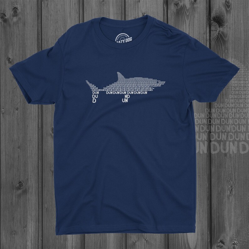Puede incluir: Una camiseta azul marino con una silueta de tibur&oacute;n blanca formada por la repetici&oacute;n de la palabra "dun". La camiseta tiene un peque&ntilde;o logotipo en la esquina superior izquierda que dice "AZY DOG".