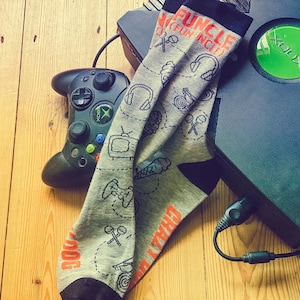 May include: A black Xbox console with a green power button and a black controller. A gray sock with a funcle design is lying on top of the console. The sock has a variety of images on it, including a joystick, a headset, and a TV.