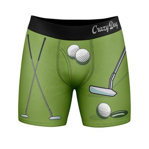 Puede incluir: Calzoncillos tipo bóxer verdes con una cintura negra que lleva el texto Crazy Dog. La tela presenta un estampado de temática de golf con palos de golf cruzados, palos individuales, pelotas de golf blancas y un hoyo oscuro.
