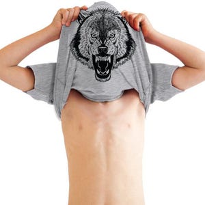 Puede incluir: Una camiseta de manga corta en color gris jaspeado que presenta una ilustración gráfica detallada en blanco y negro de una cabeza de lobo gruñendo con los dientes al descubierto y ojos intensos, centrada en la parte frontal de la prenda.