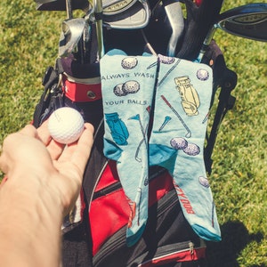 May include: A blue and white golf-themed sock with the text "Always Wash Your Balls" and a golf bag design. A golf bag with golf clubs and a white golf ball are in the foreground.