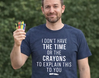 May include: A navy blue short-sleeved t-shirt featuring the white printed text I DON'T HAVE THE TIME OR THE CRAYONS TO EXPLAIN THIS TO YOU, accompanied by a small graphic of a broken crayon at the bottom, held alongside a small cluster of crayons.