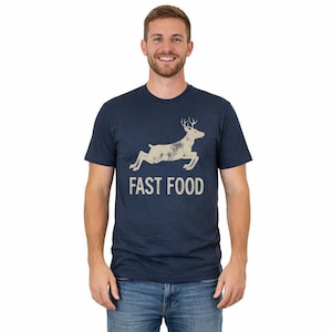 May include: A navy blue short-sleeved t-shirt featuring a distressed beige graphic of a leaping deer above the text "FAST FOOD" in a bold, sans-serif font. The shirt has a crew neckline and a standard fit, constructed from a soft fabric.