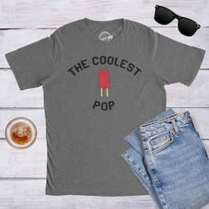 May include: A heather gray short-sleeve t-shirt featuring the black text THE COOLEST POP arched over a graphic of a red popsicle on a wooden stick. The shirt is accompanied by a pair of blue denim jeans and black sunglasses.