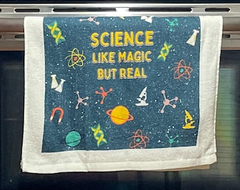 May include: A rectangular kitchen towel with a dark blue background featuring a white speckled pattern and various science-themed icons including DNA strands, planets, atoms, and microscopes. The text reads: SCIENCE LIKE MAGIC BUT REAL.