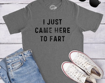 Op de afbeelding: Een grijs gemêleerd T-shirt met korte mouwen en de zwarte tekst I JUST CAME HERE TO FART op de borst, vergezeld door een blauwe spijkerbroek, witte canvas sneakers en een zwarte zonnebril op een witte houten ondergrond.