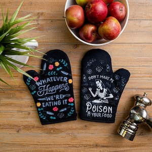 May include: A pair of black oven mitts featuring white and colorful graphic designs. One mitt displays the text "WHATEVER HAPPENS WE'RE EATING IT" with floral and knife motifs. The other mitt shows a person with a pie and the text "DON'T MAKE ME POISON YOUR FOOD".