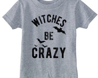 Könnte beinhalten: Ein graues, meliertes Kurzarm-T-Shirt mit dem schwarzen, abgenutzten Schriftzug WITCHES BE CRAZY auf der Vorderseite. Das Design enthält drei schwarze Silhouetten von fliegenden Fledermäusen, die um die Großbuchstaben verteilt sind.