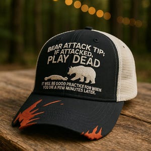 May include: Black and tan trucker hat with the text "BEAR ATTACK TIP; IF ATTACKED, PLAY DEAD" and a bear graphic. The hat also includes the text "IT WILL BE GOOD PRACTICE FOR WHEN YOU DIE A FEW MINUTES LATER." Orange flame accents are on the brim.