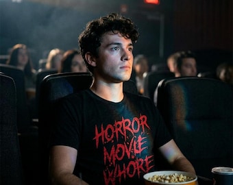 May include: A black short-sleeved t-shirt featuring the red text HORROR MOVIE WHORE printed on the front. The shirt is worn by a person sitting in a dark cinema, holding a yellow bucket of popcorn with white text and graphics.