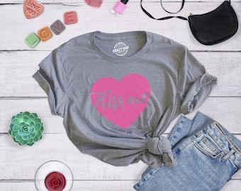 Könnte beinhalten: Ein graues, meliertes Kurzarm-T-Shirt mit einem großen rosa Herz-Aufdruck auf der Brust, in dessen Mitte der Schriftzug "Kiss me" in Schreibschrift steht. Das T-Shirt hat einen Rundhalsausschnitt und wird zusammen mit einer blauen Jeans gezeigt.